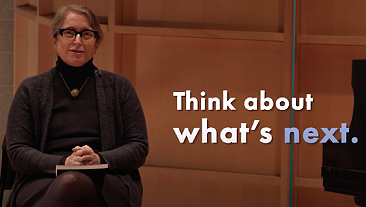 Provost Linda Essigs sits on a stage, with on screen text reading "Think about what's next."