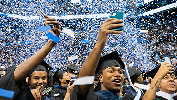 Three Baruch College students celebrate graduation, in full academic regalia, at Barclays Center. They are smiling and cheering witht heir hands in the air. COnfetti is falling in the background.