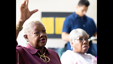 An elderly woman raises her hand in a group discussion setting.