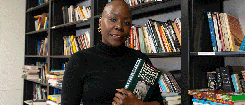 A Baruch College professor holds a book titled Frederick Douglass standing in front of several bookshelves.